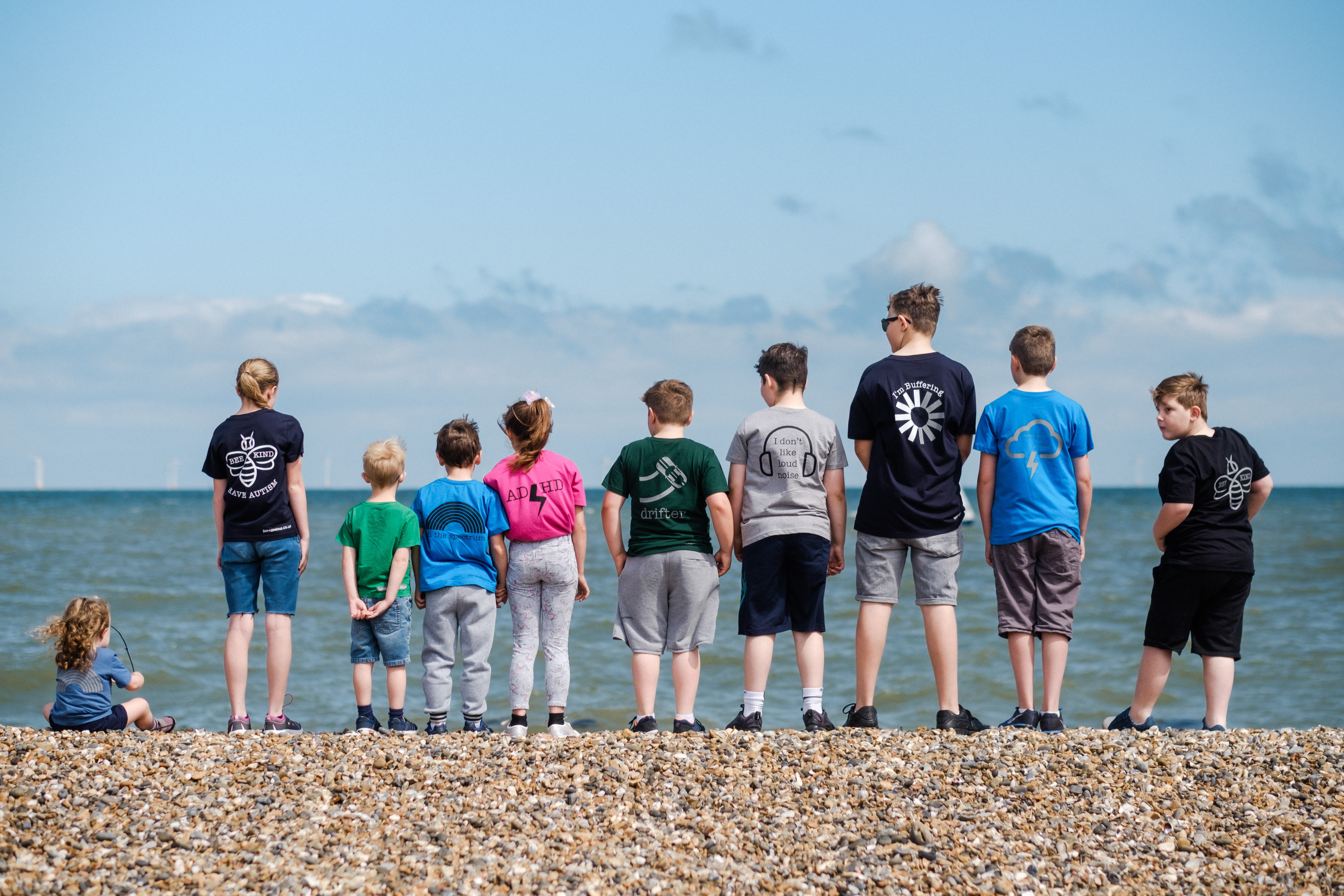 A group of children wear Born Anxious clothing standing on the beach looking out to sea